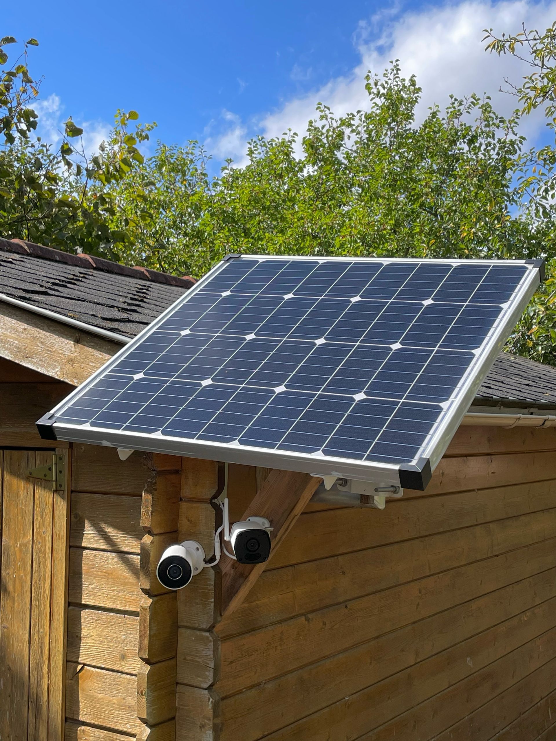 Panneau solaire installé sur le toit d'un abri en bois, à côté de deux caméras de sécurité, sous un ciel bleu.