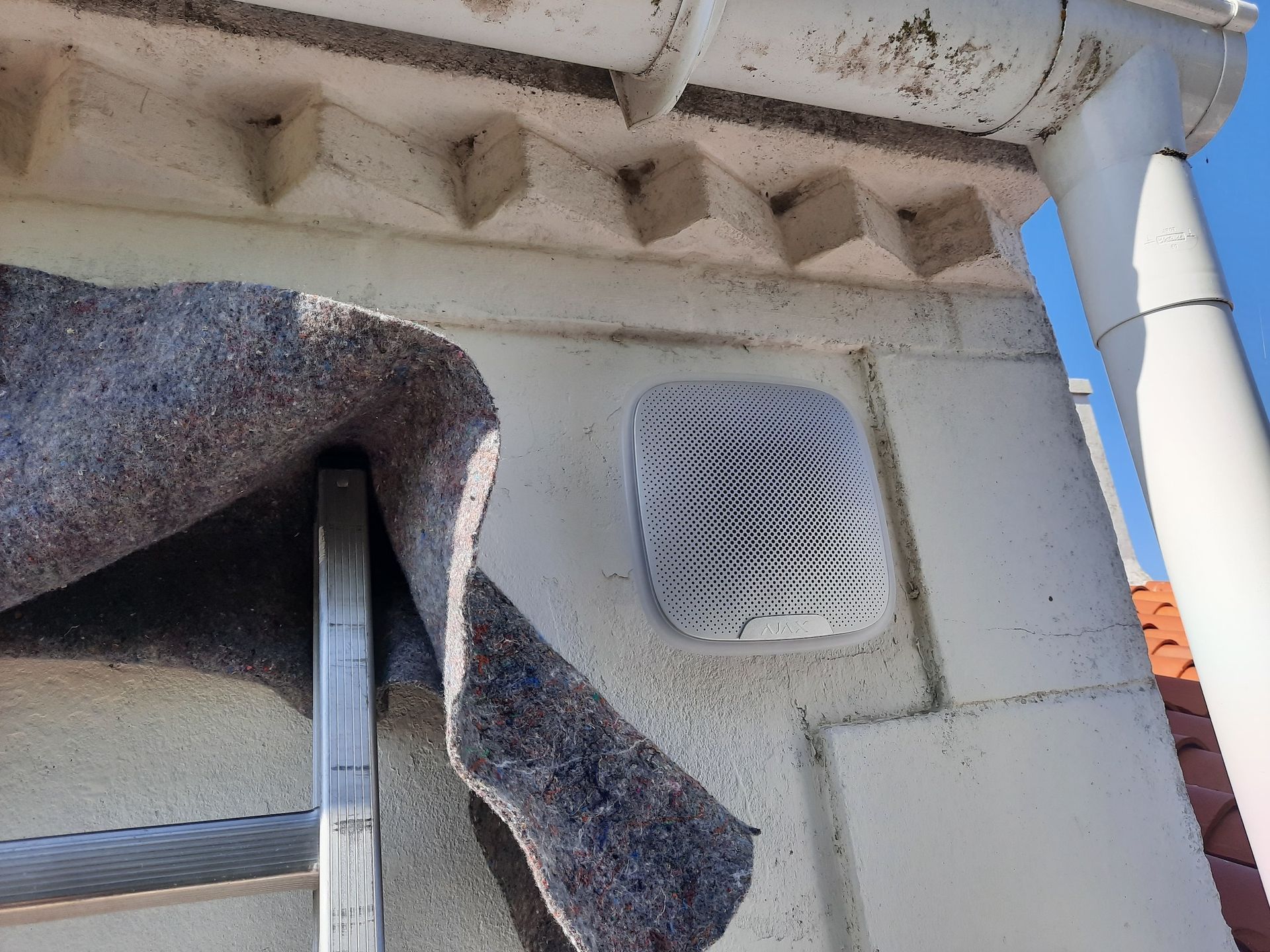 Gros plan sur l'extérieur d'un bâtiment montrant des ouvertures de ventilation, des canalisations et un morceau de tissu sombre drapé sur un ciel bleu.