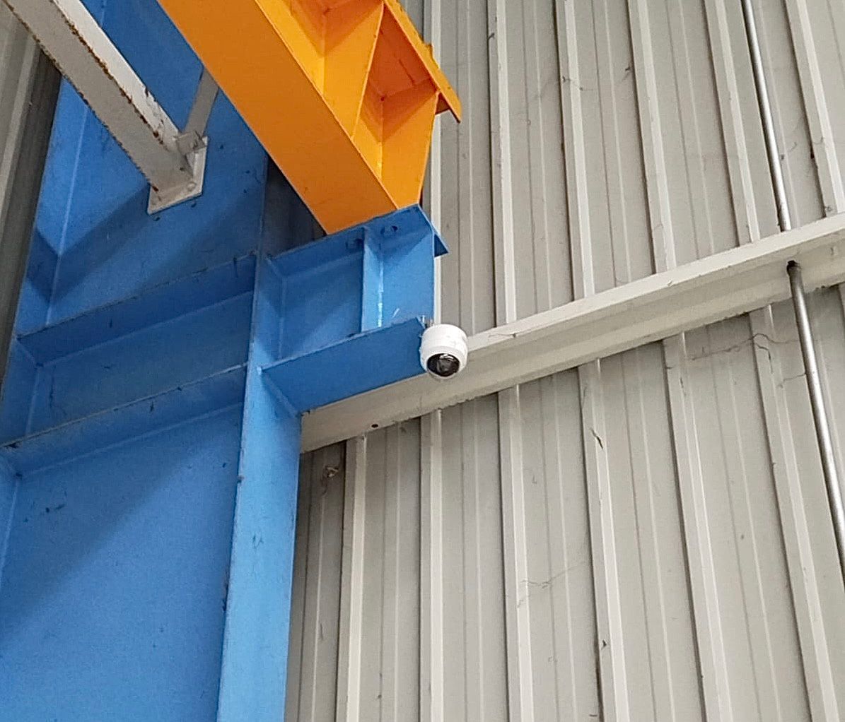 Structure industrielle bleue avec poutre orange et caméra de sécurité blanche fixée sur un mur ondulé gris