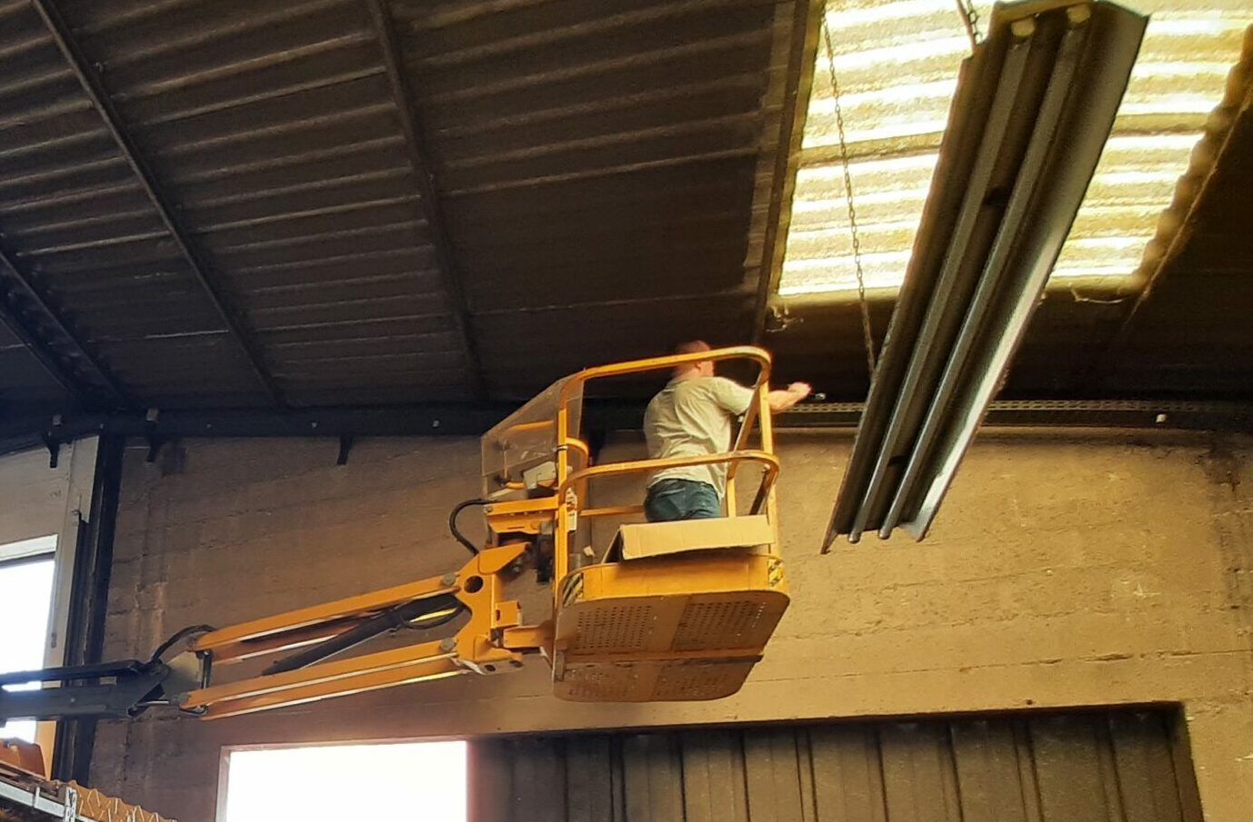 Un ouvrier sur une nacelle élévatrice jaune à l'intérieur d'un entrepôt, tendant le bras presque jusqu'au plafond.