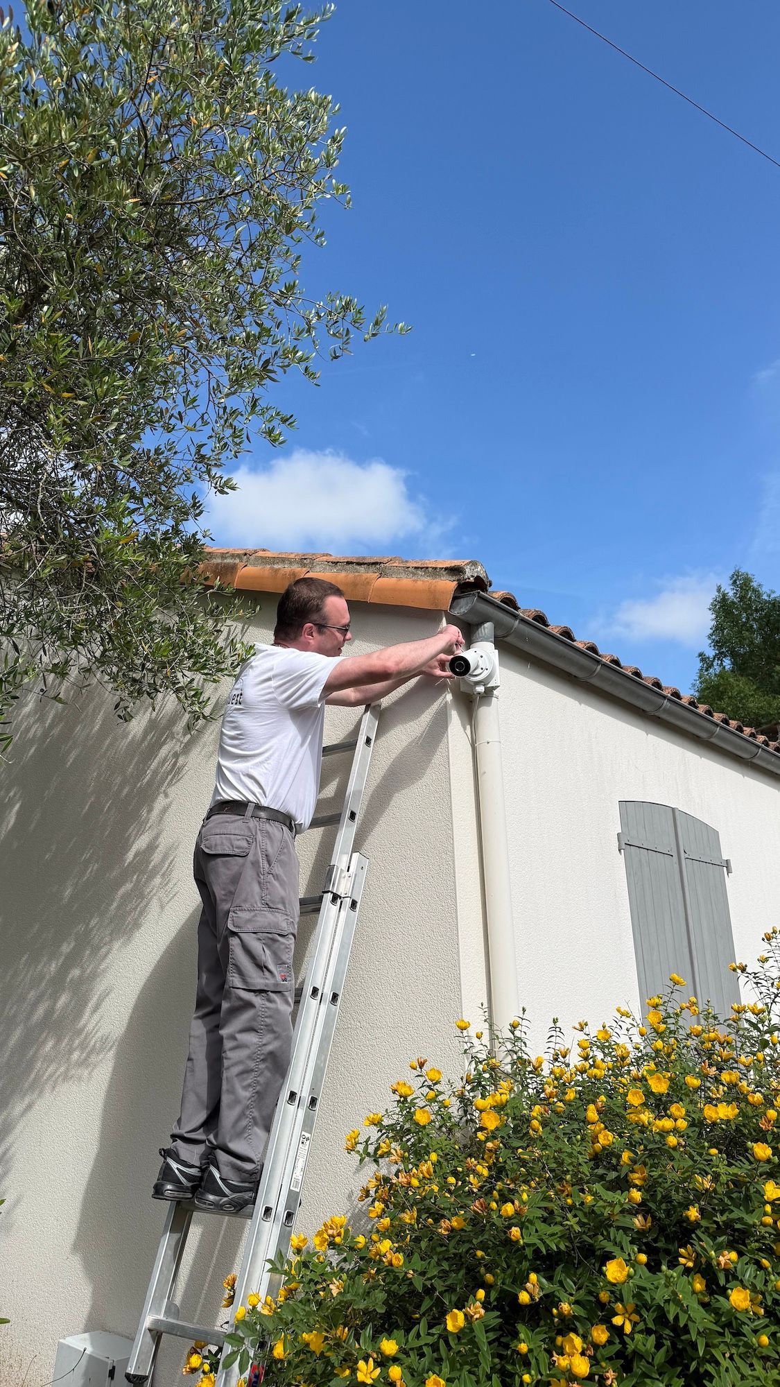 Une personne, debout sur une échelle, nettoie une gouttière de toit à côté d'une maison blanche et de fleurs jaunes.