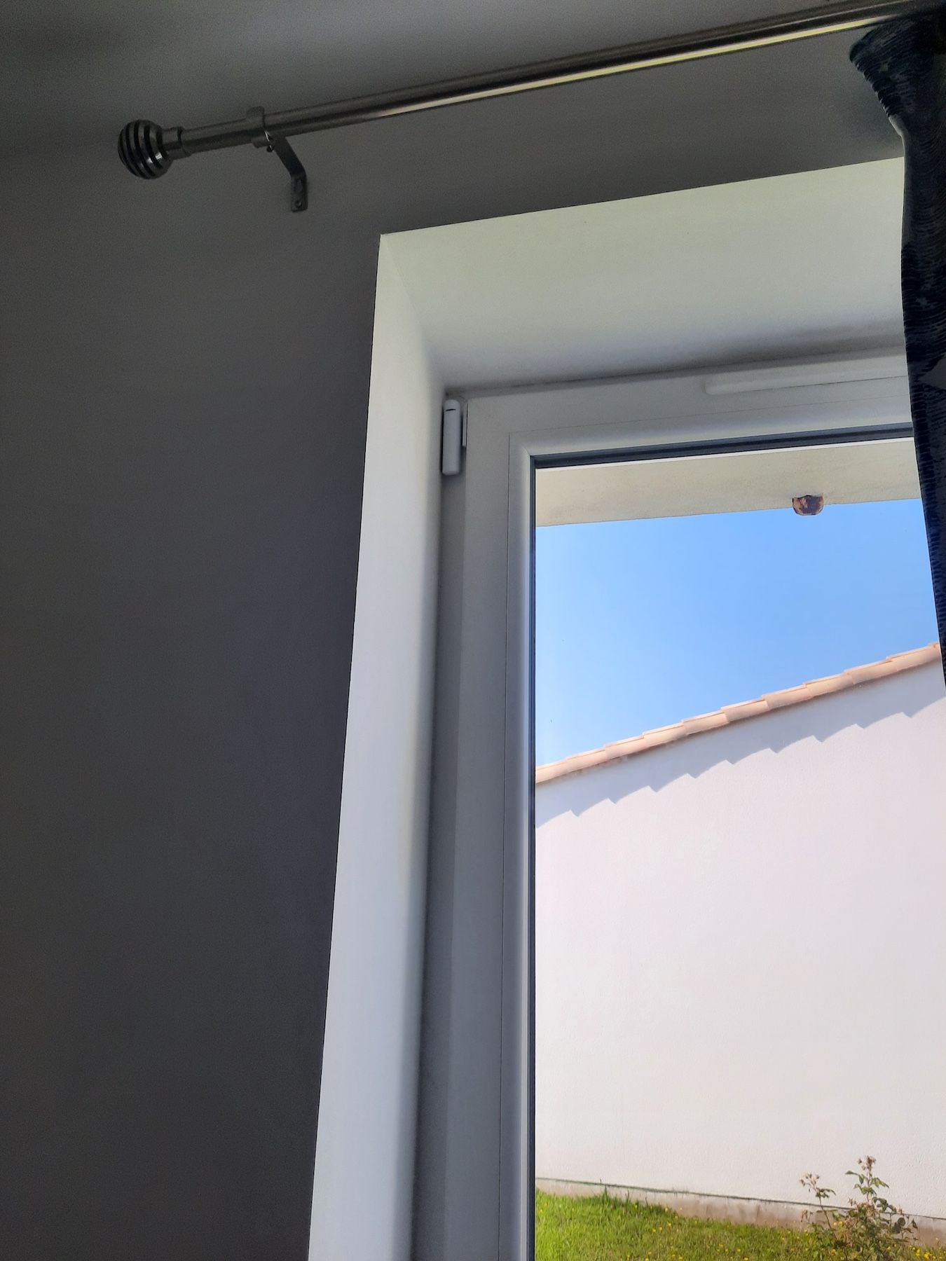 Mur gris et fenêtre avec tringle à rideaux noire, donnant sur un ciel bleu et un bâtiment blanc à l'extérieur.
