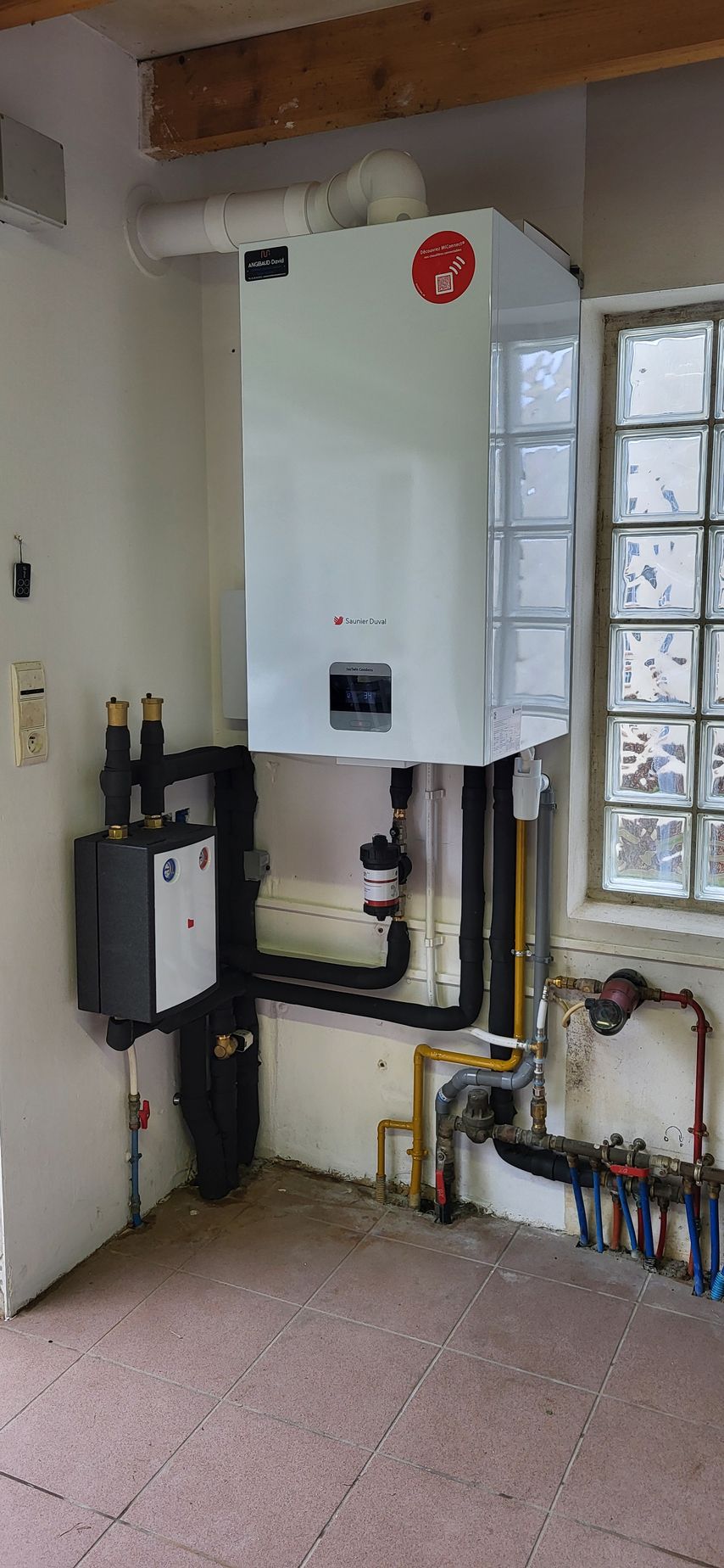 Chaudière murale blanche avec plomberie, près d'une fenêtre à vitres en pavés de verre.