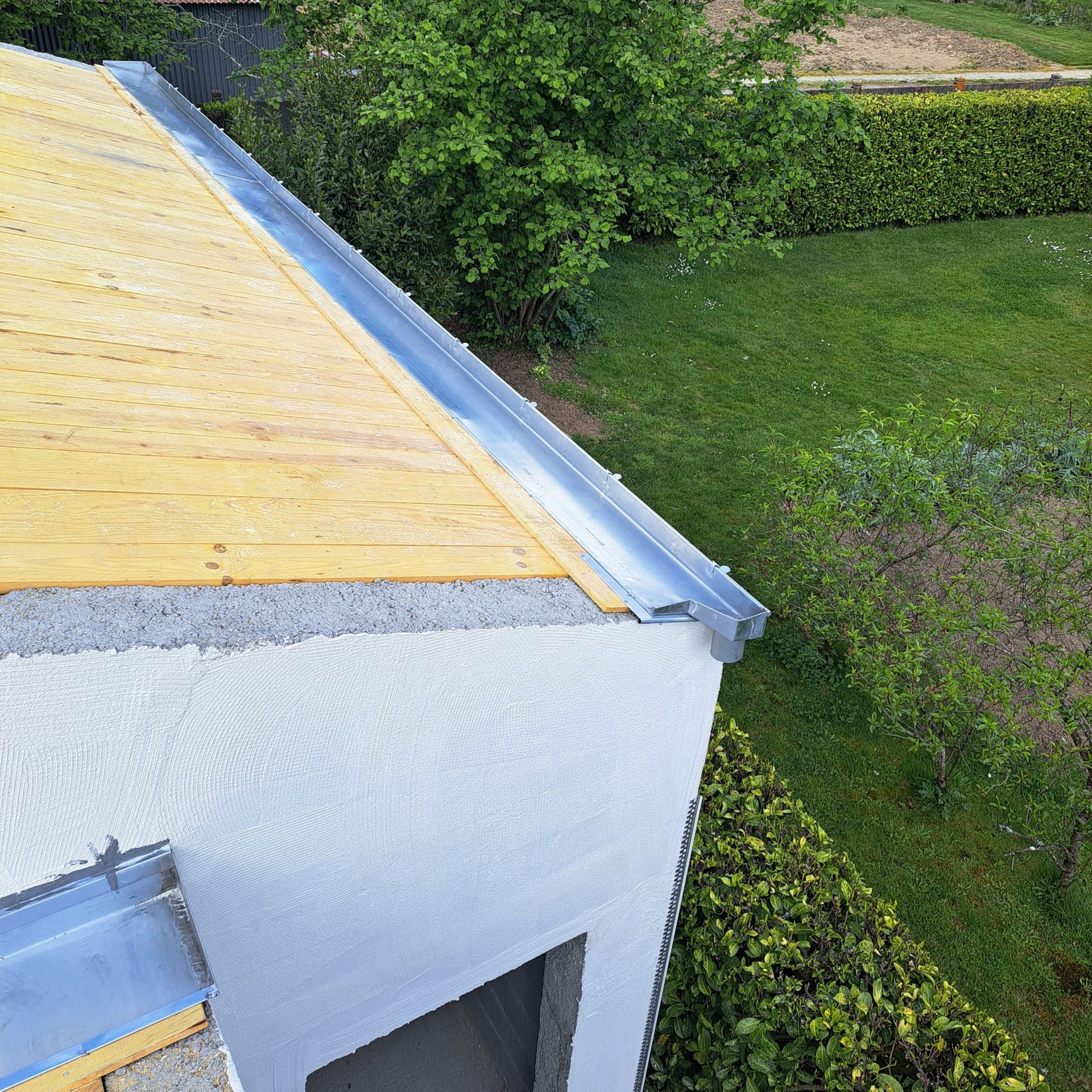 Bord de toit avec une gouttière en zinc et surface en bois brut, jouxtant un mur en stuc blanc et une cour verdoyante.