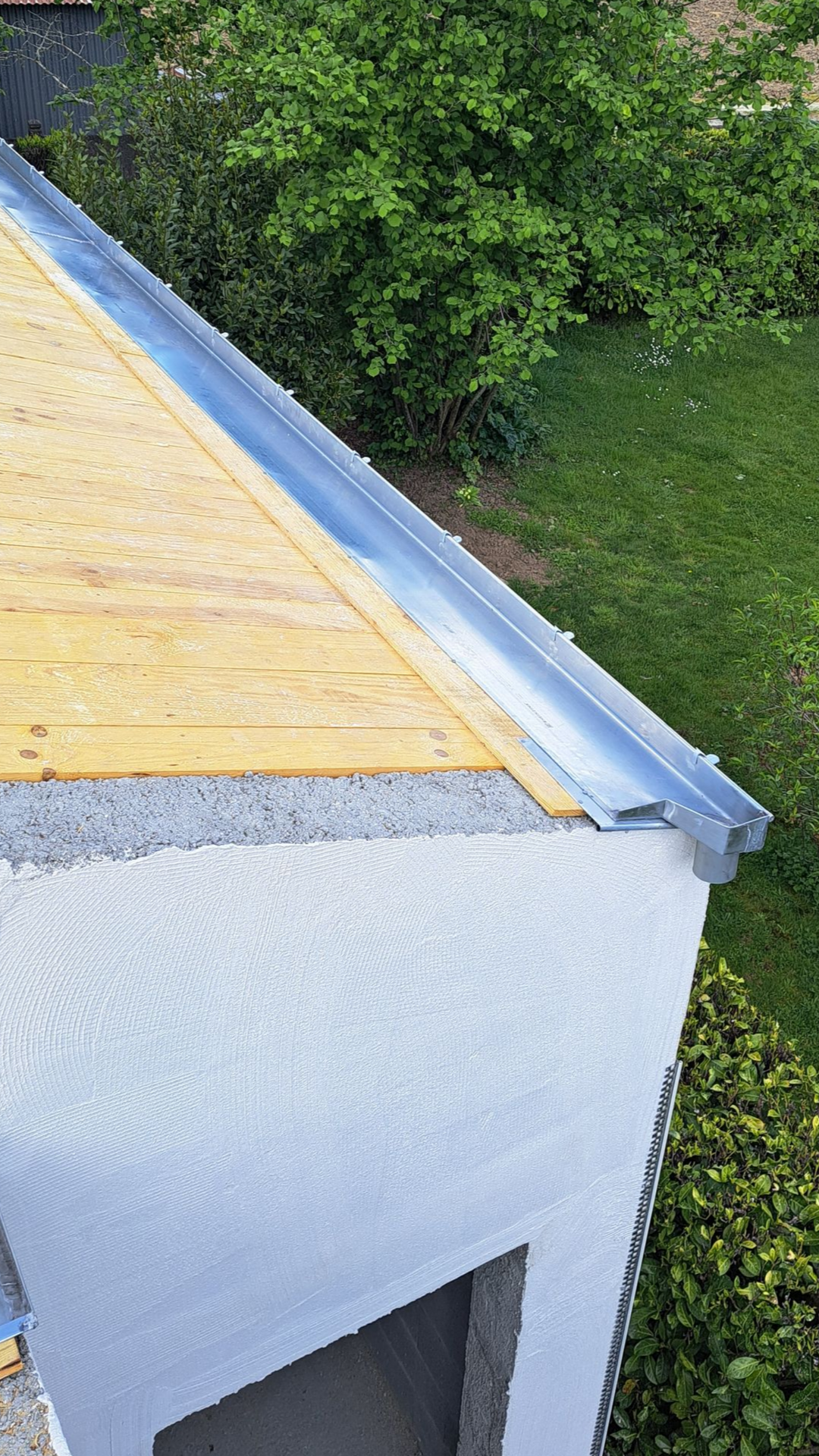 Bord de toit avec gouttière en zinc et revêtement en bois, à côté d'un mur en stuc blanc et de feuillage vert.