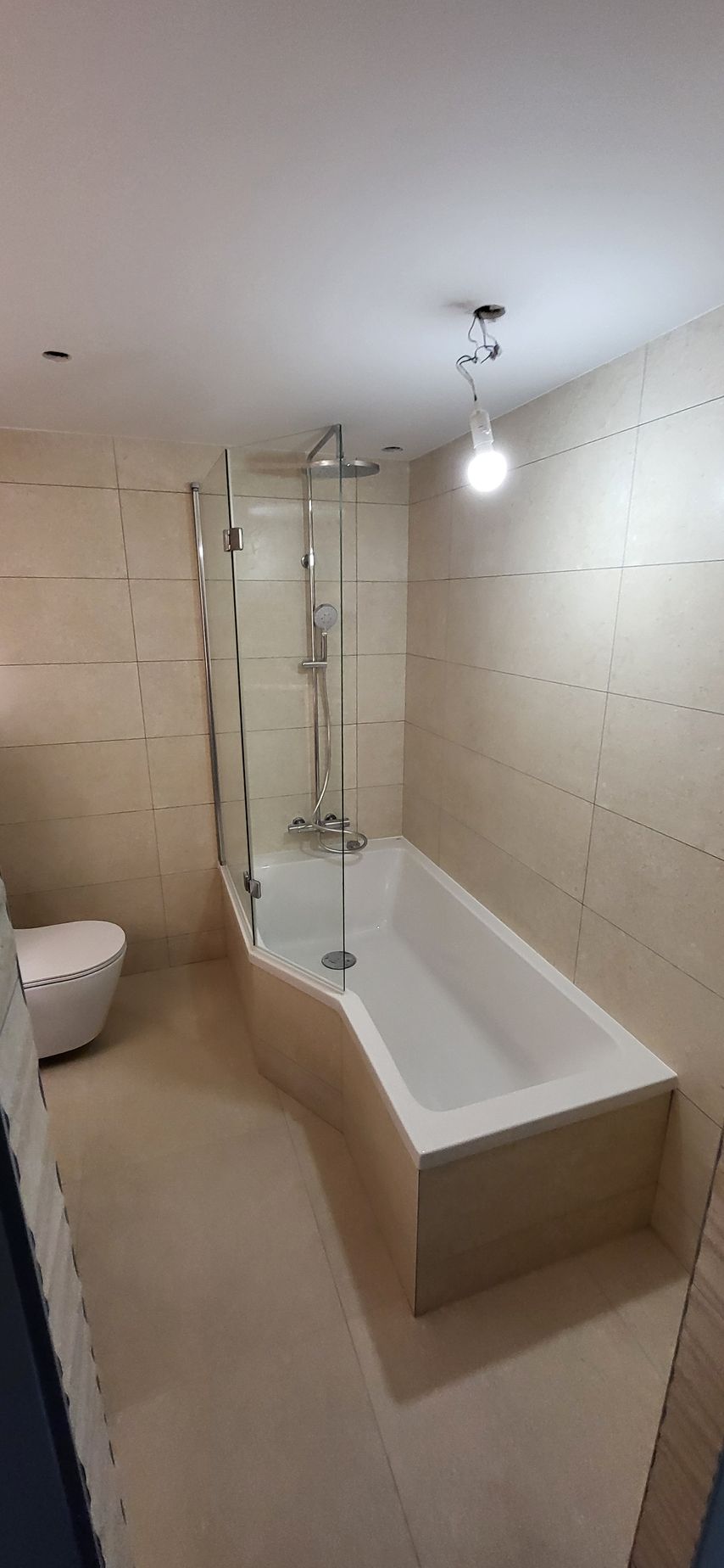 Salle de bains avec baignoire blanche, douche et toilettes ; murs carrelés beiges.