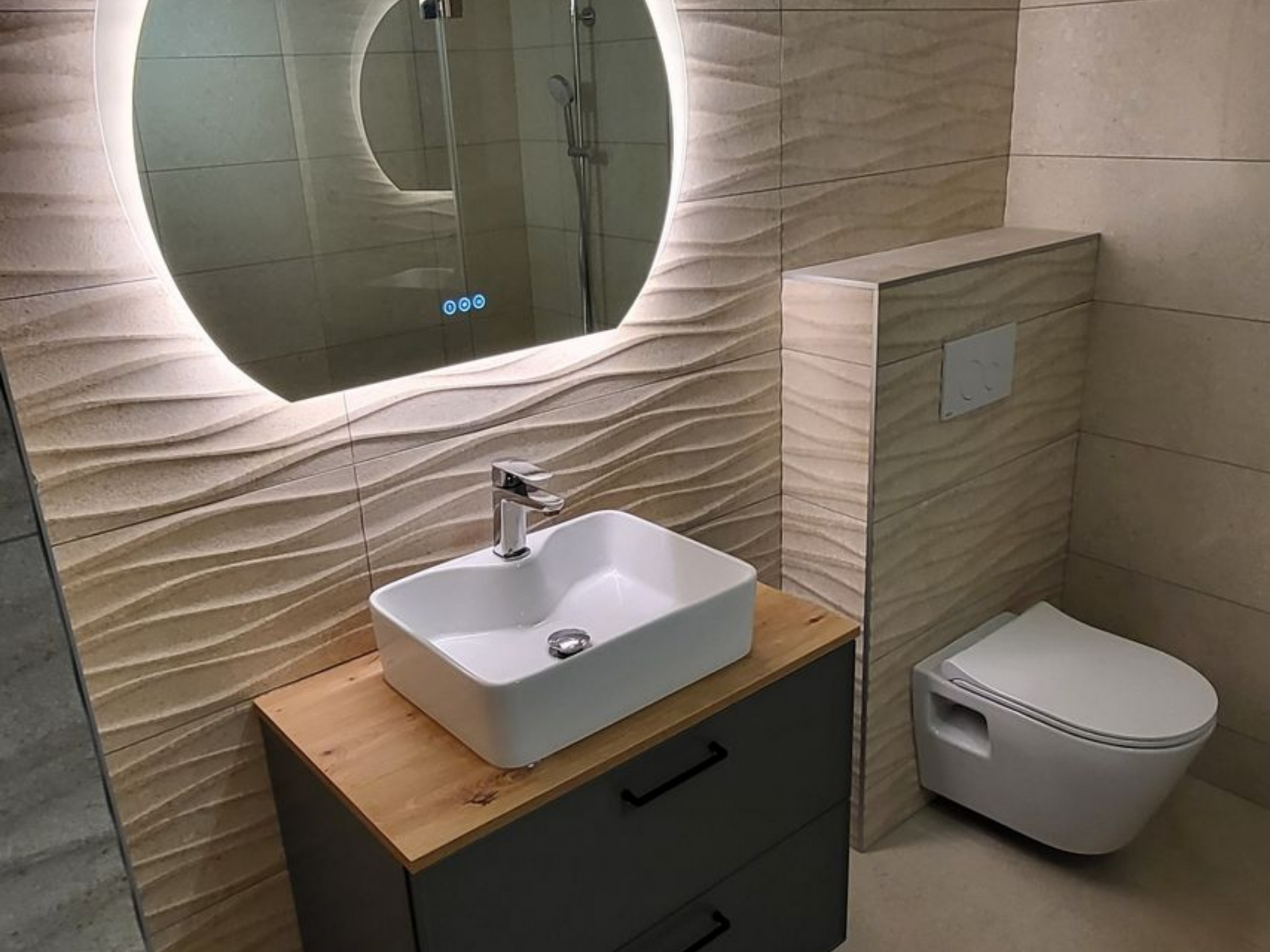 Salle de bains moderne avec carrelage beige ondulé, miroir rond éclairé, toilettes suspendues et lavabo sur meuble gris.