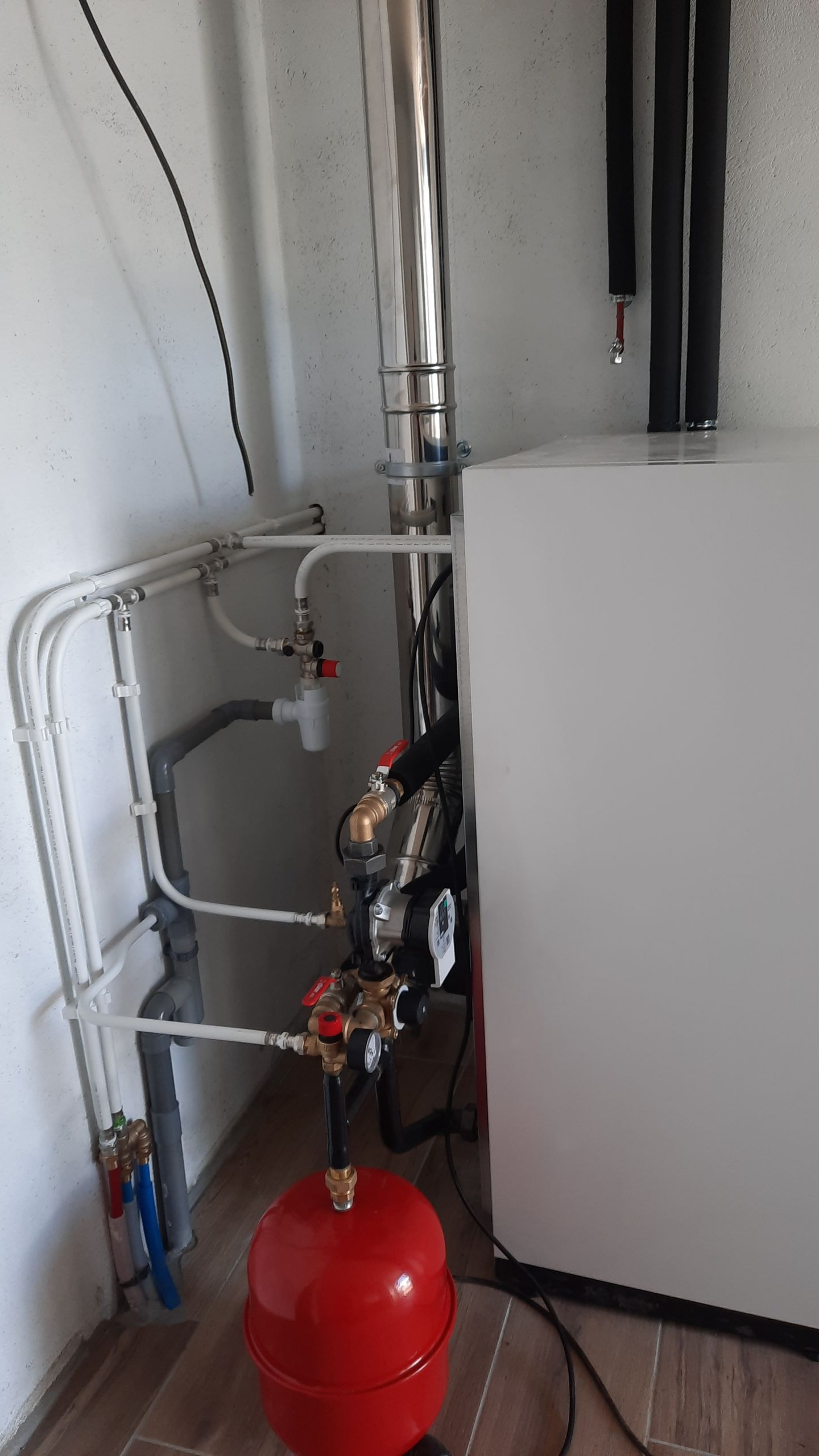 Composants du système de chauffage avec tuyaux, un réservoir rouge et une unité blanche dans une buanderie.