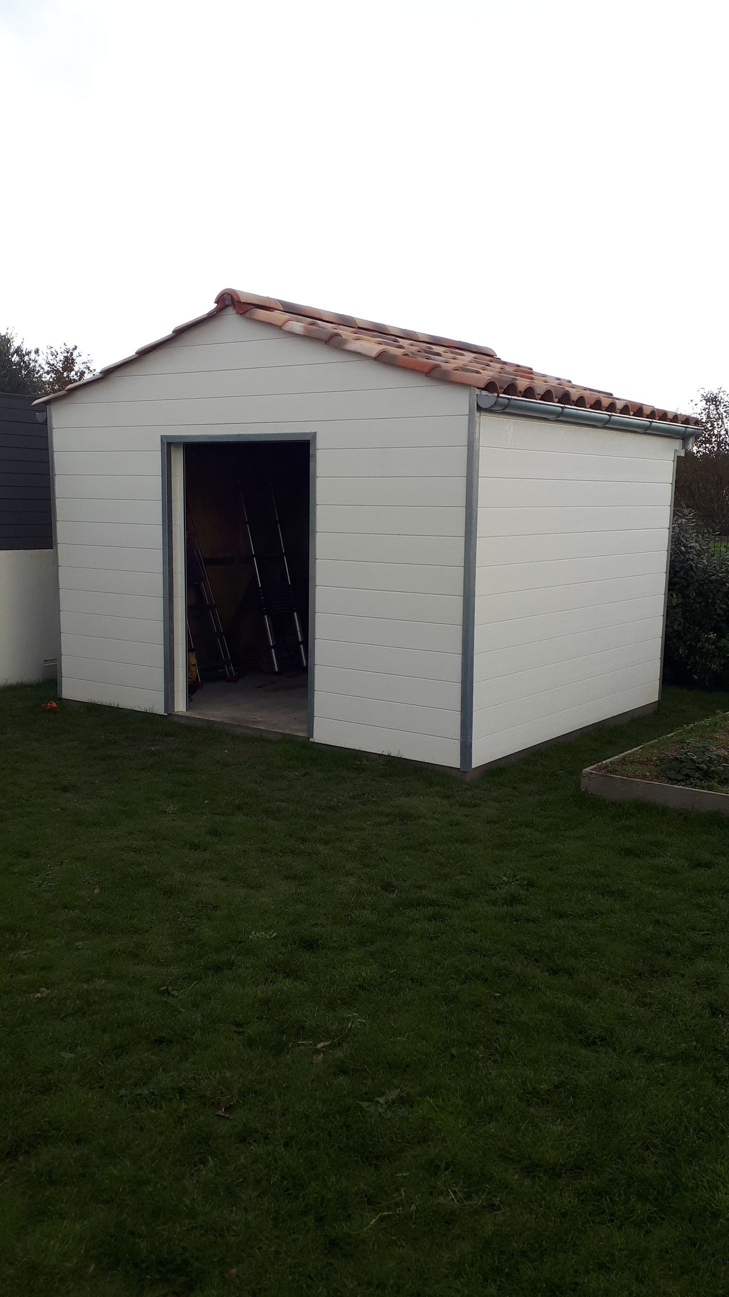 Abri de jardin blanc avec porte ouverte, toit de tuiles rouges et pelouse verte.