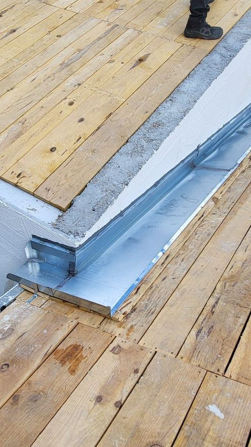 Des gouttières en zinc sont installées entre les planches d'une terrasse en bois, avec du béton et une jambe de personne en arrière-plan.