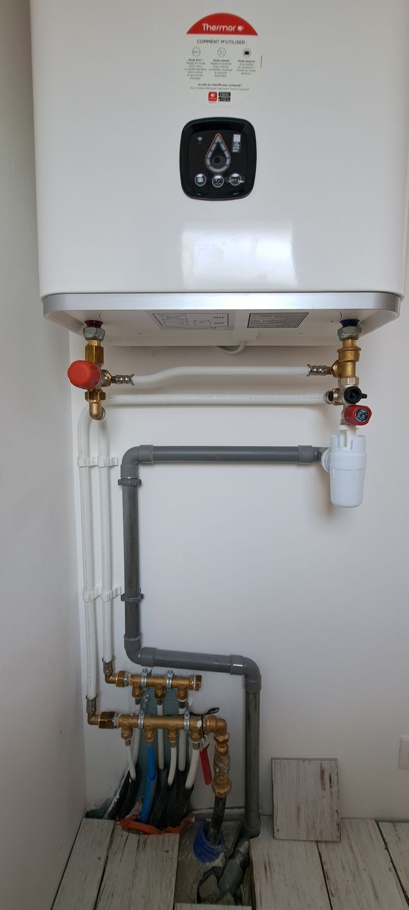 Un chauffe-eau blanc avec ses tuyaux et vannes raccordés, dans un coin de la pièce.
