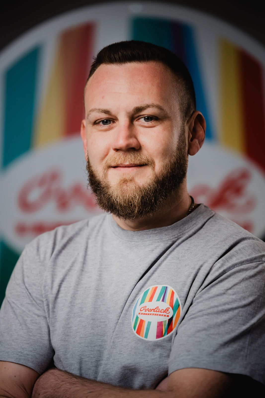 Ein Mann mit Bart steht mit verschränkten Armen vor einem bunten Schild.