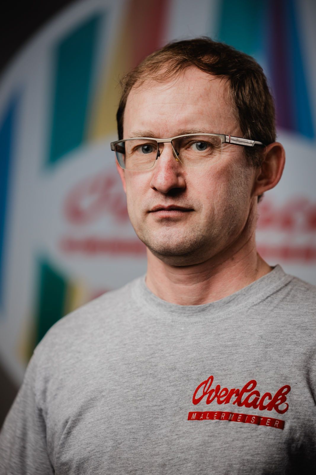 Ein Mann mit Brille und grauem Hemd steht vor einem bunten Hintergrund.