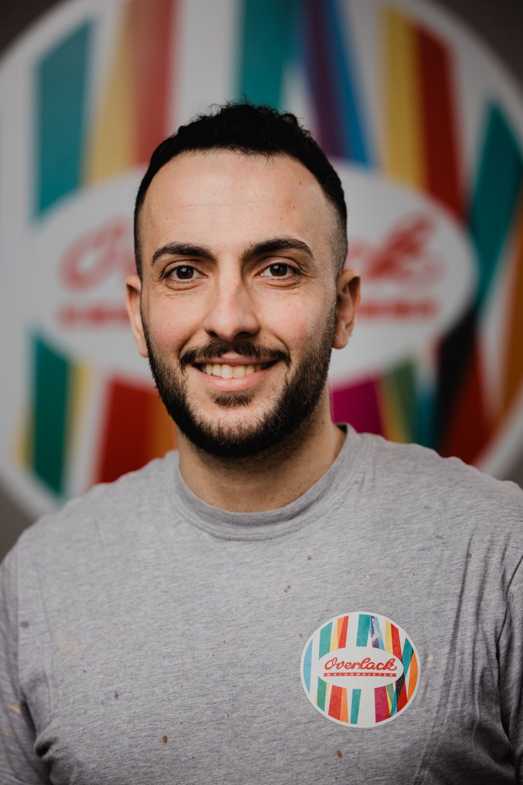 Ein Mann mit Bart lächelt vor einem bunten Schild.