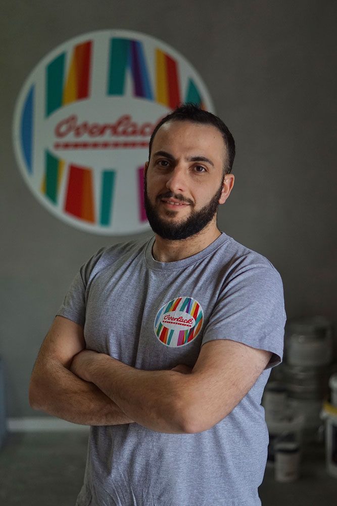 Ein Mann mit verschränkten Armen in einem grauen T-Shirt steht vor einem farbenfrohen, gestreiften Logo an einer Wand.