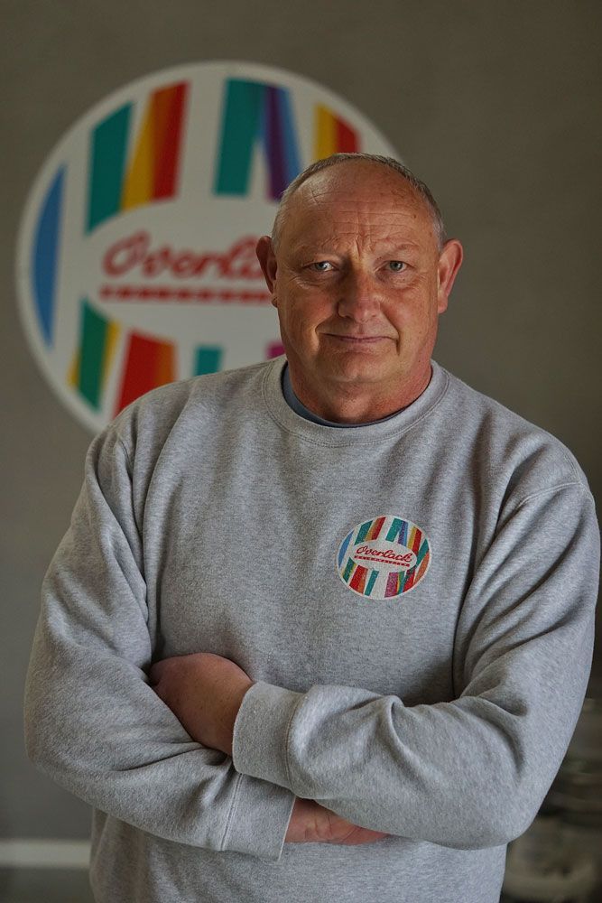 Ein Mann in einem grauen Sweatshirt steht mit verschränkten Armen vor einer farbenfrohen, gestreiften Logowand.