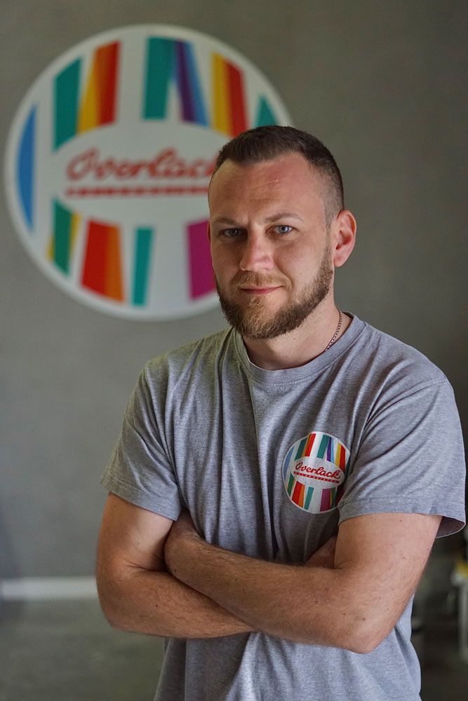 Ein Mann mit verschränkten Armen steht in einem grauen T-Shirt vor einem farbenfrohen, gestreiften Logo-Hintergrund.