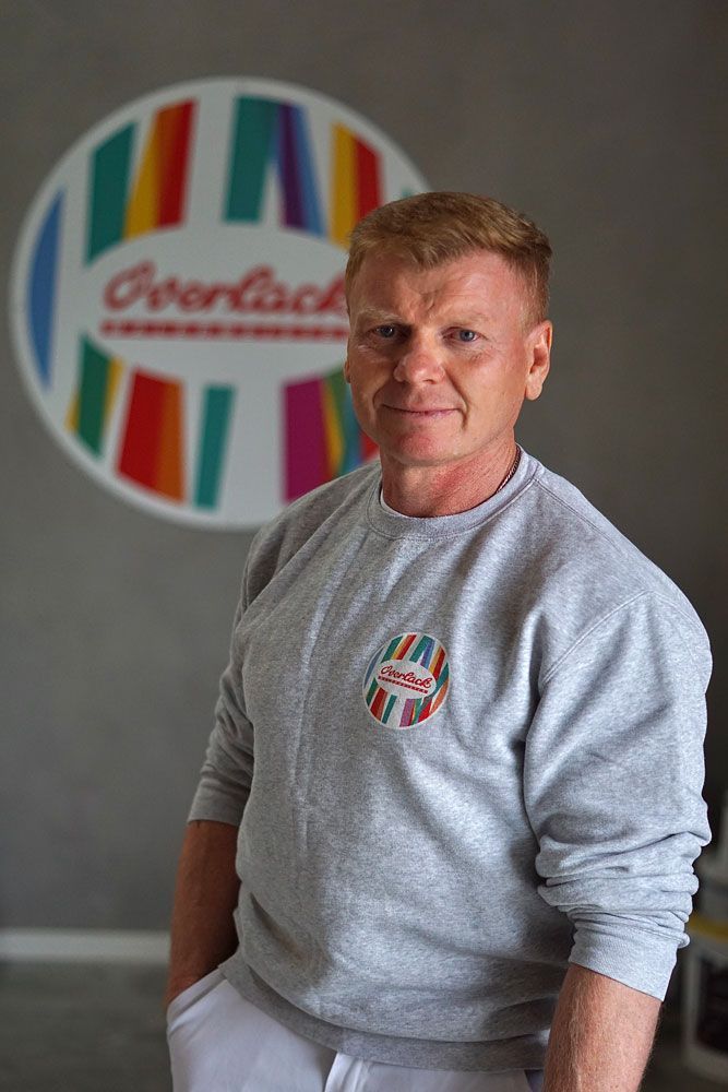 Ein Mann in einem grauen Sweatshirt steht drinnen vor einem farbenfrohen, runden Logo-Wandschild.
