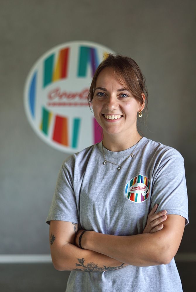 Eine lächelnde Person in einem grauen T-Shirt steht mit verschränkten Armen vor einem farbenfrohen, kreisförmigen Logo.