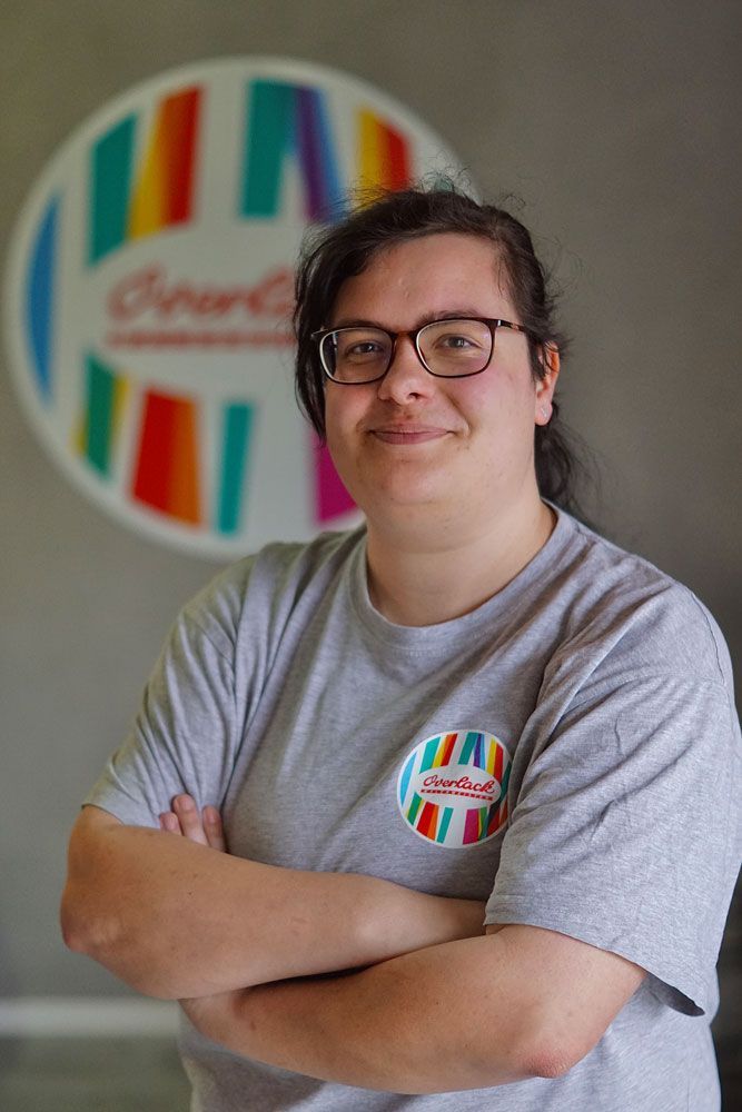Person mit Brille und grauem T-Shirt, die Arme verschränkt, steht vor einem farbenfrohen, kreisförmigen Logo.