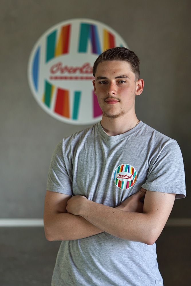 Junger Mensch in einem grauen T-Shirt, die Arme verschränkt, steht vor einem bunten, runden Logo an einer grauen Wand