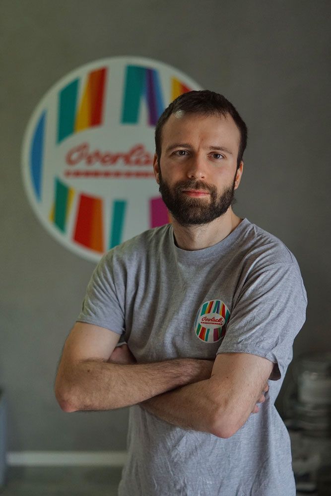 Mann mit verschränkten Armen in einem grauen T-Shirt, der vor einem bunten, kreisförmigen Logo an einer Wand steht