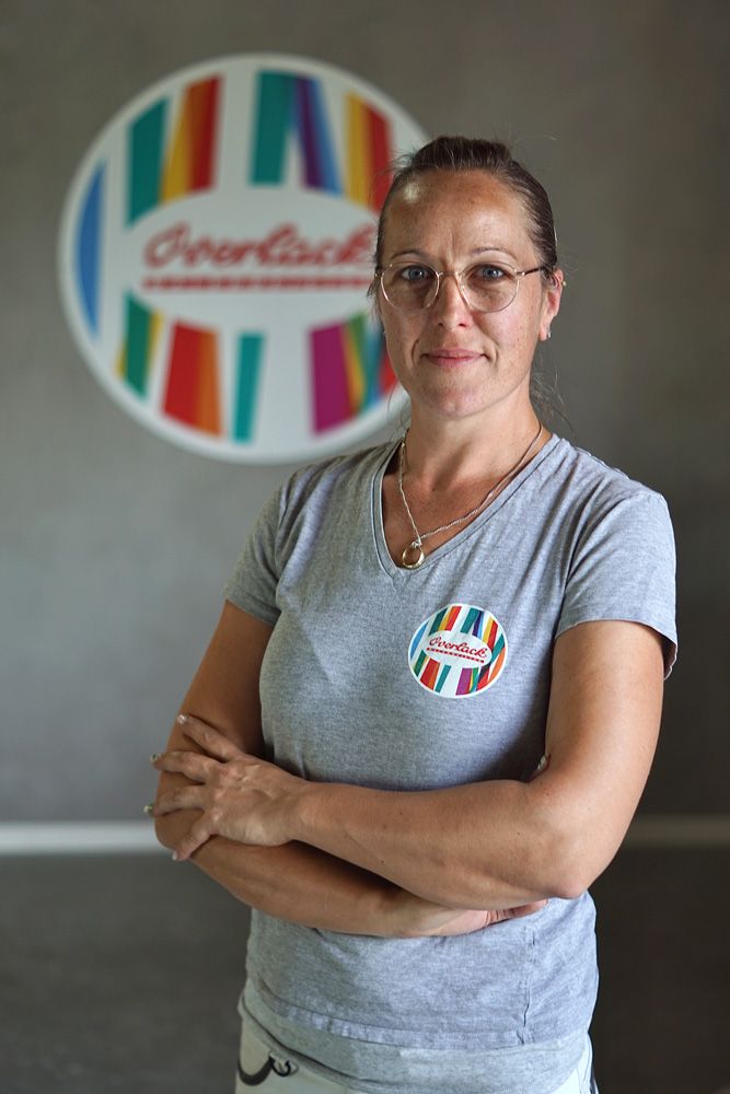 Frau in einem grauen T-Shirt steht mit verschränkten Armen vor einem farbenfrohen Logo an einer Wand.