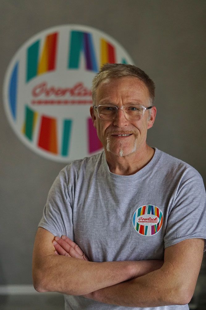 Ein Mann mit verschränkten Armen steht vor einem farbenfrohen, runden Logo. Er trägt ein graues Hemd und eine Brille.