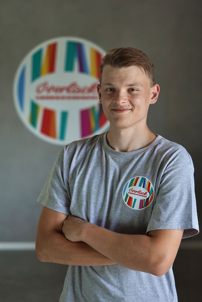 Eine Person mit verschränkten Armen in einem grauen T-Shirt steht vor einem farbenfrohen, kreisförmigen Logo.