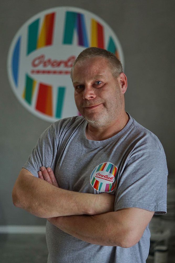 Ein Mann mit verschränkten Armen in einem grauen T-Shirt steht vor einem farbenfrohen, kreisförmigen Logo.