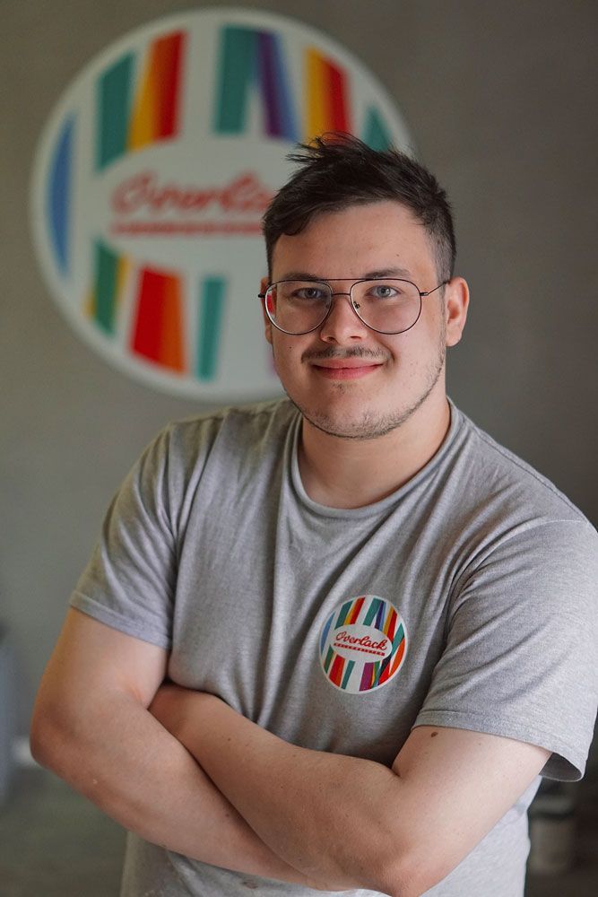 Mann mit Brille und verschränkten Armen, bekleidet mit einem grauen T-Shirt, steht vor einem bunten runden Schild.