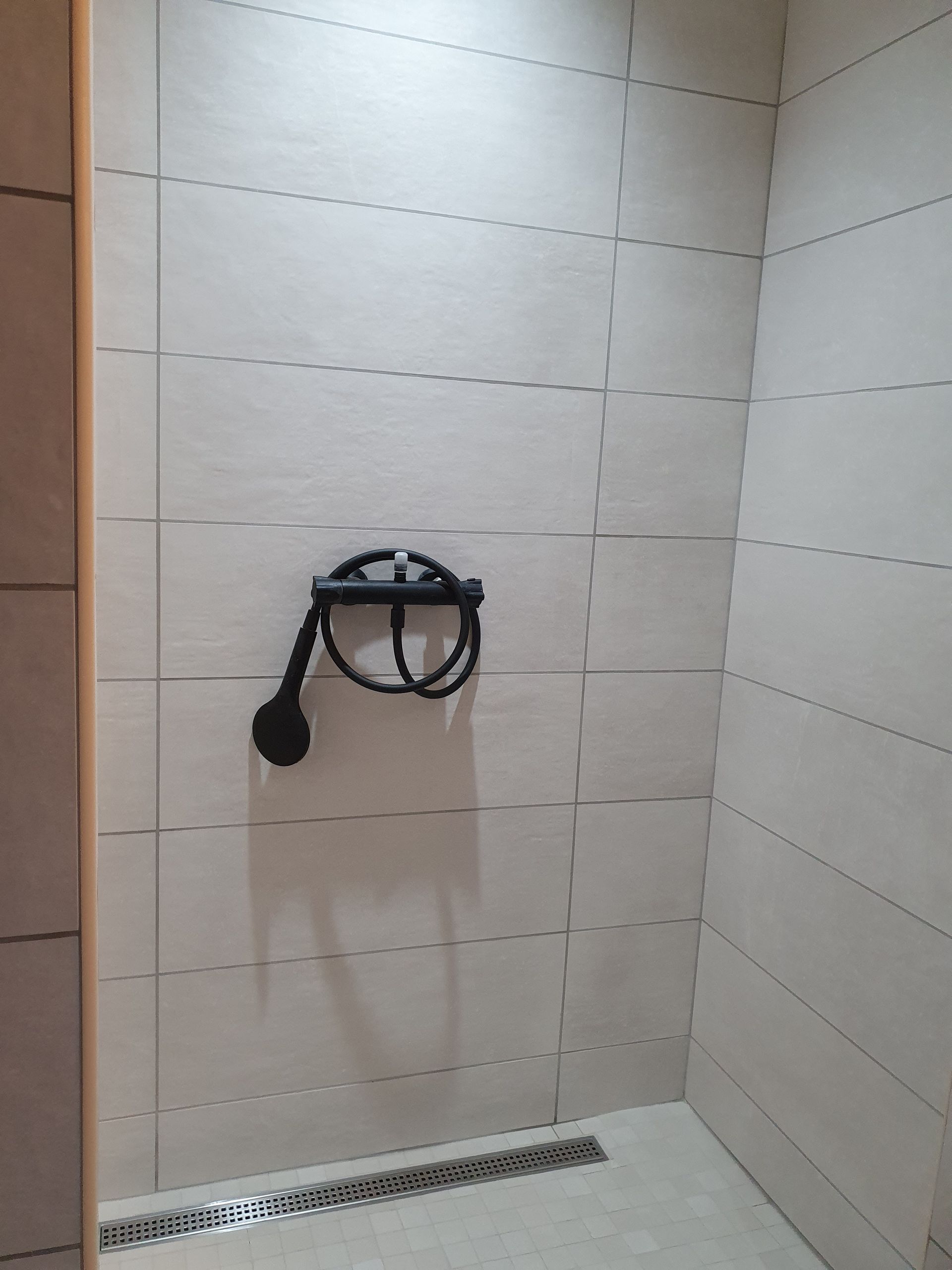 Cabine de douche avec pommeau de douche noir, fixée sur un mur carrelé de couleur claire, et caniveau linéaire.
