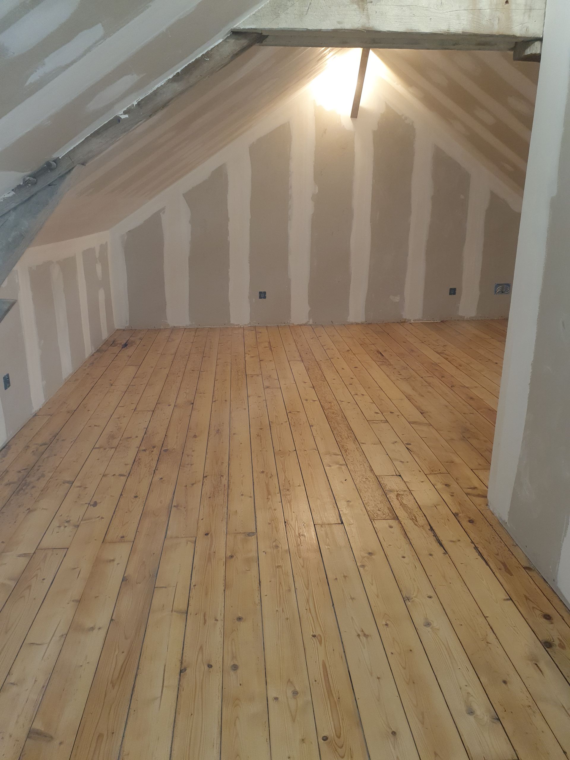 Pièce mansardée en cours d'aménagement avec plancher en bois et murs en plaques de plâtre.