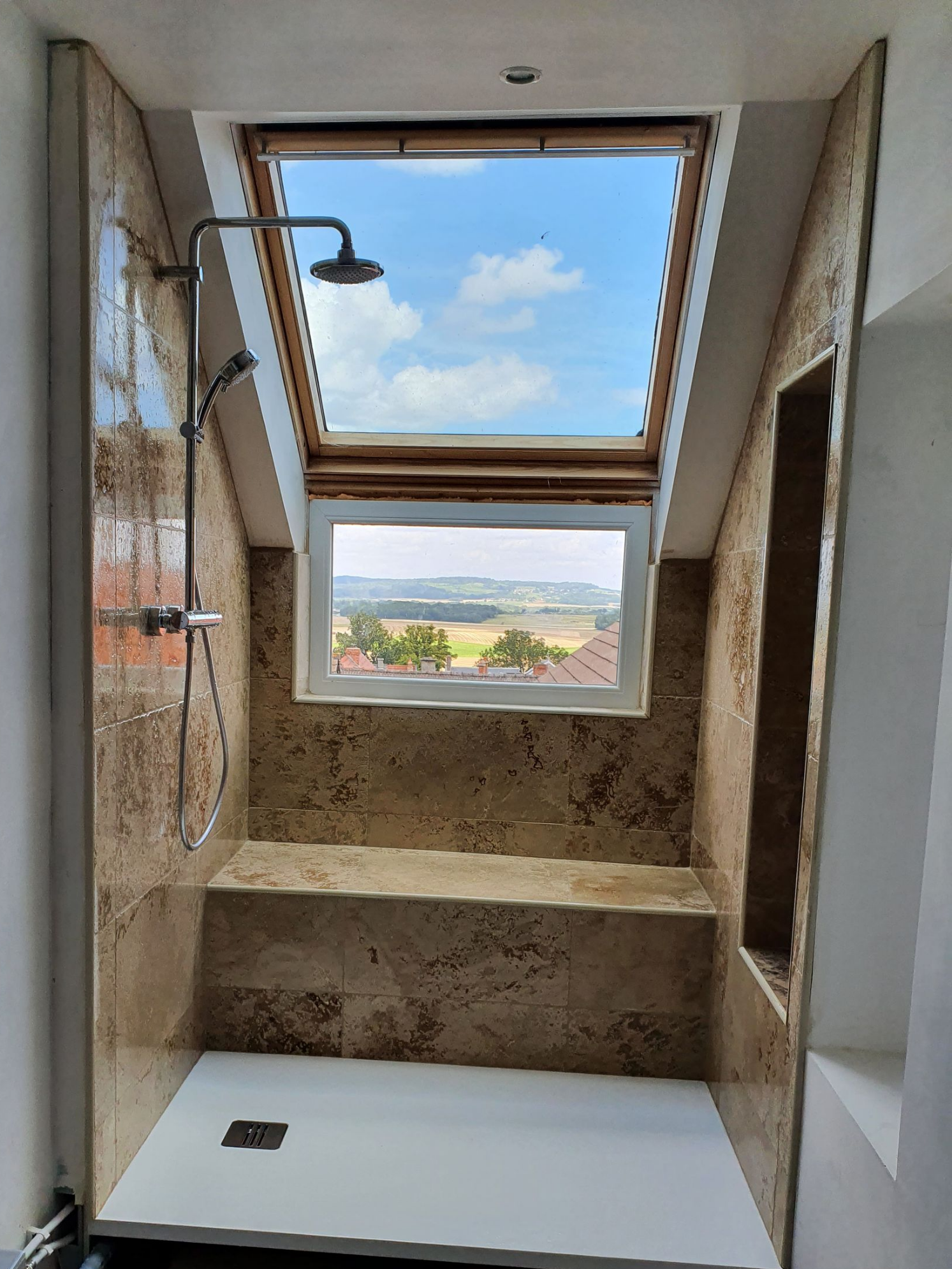 Douche avec puits de lumière et vue sur le paysage extérieur. Carrelage en pierre beige et receveur de douche blanc.