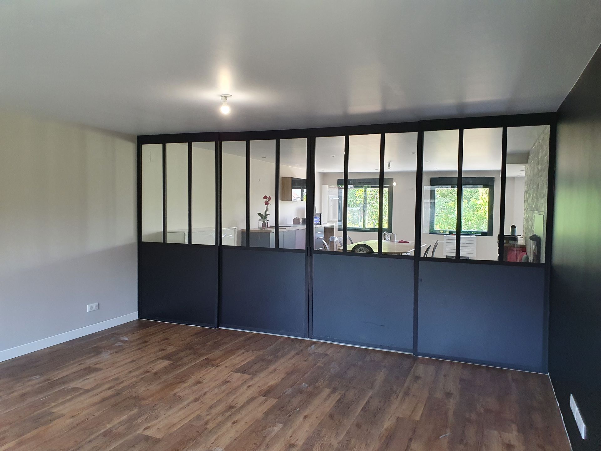 Pièce avec cloison en verre et métal noir, parquet, donnant sur une cuisine.