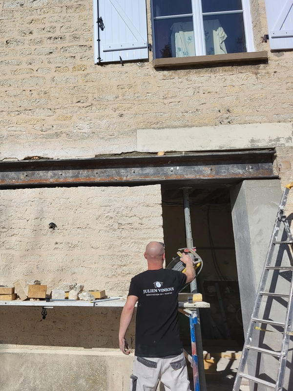 Une personne utilise un outil électrique sur un mur sous une poutre en acier, avec une fenêtre et des volets au-dessus.