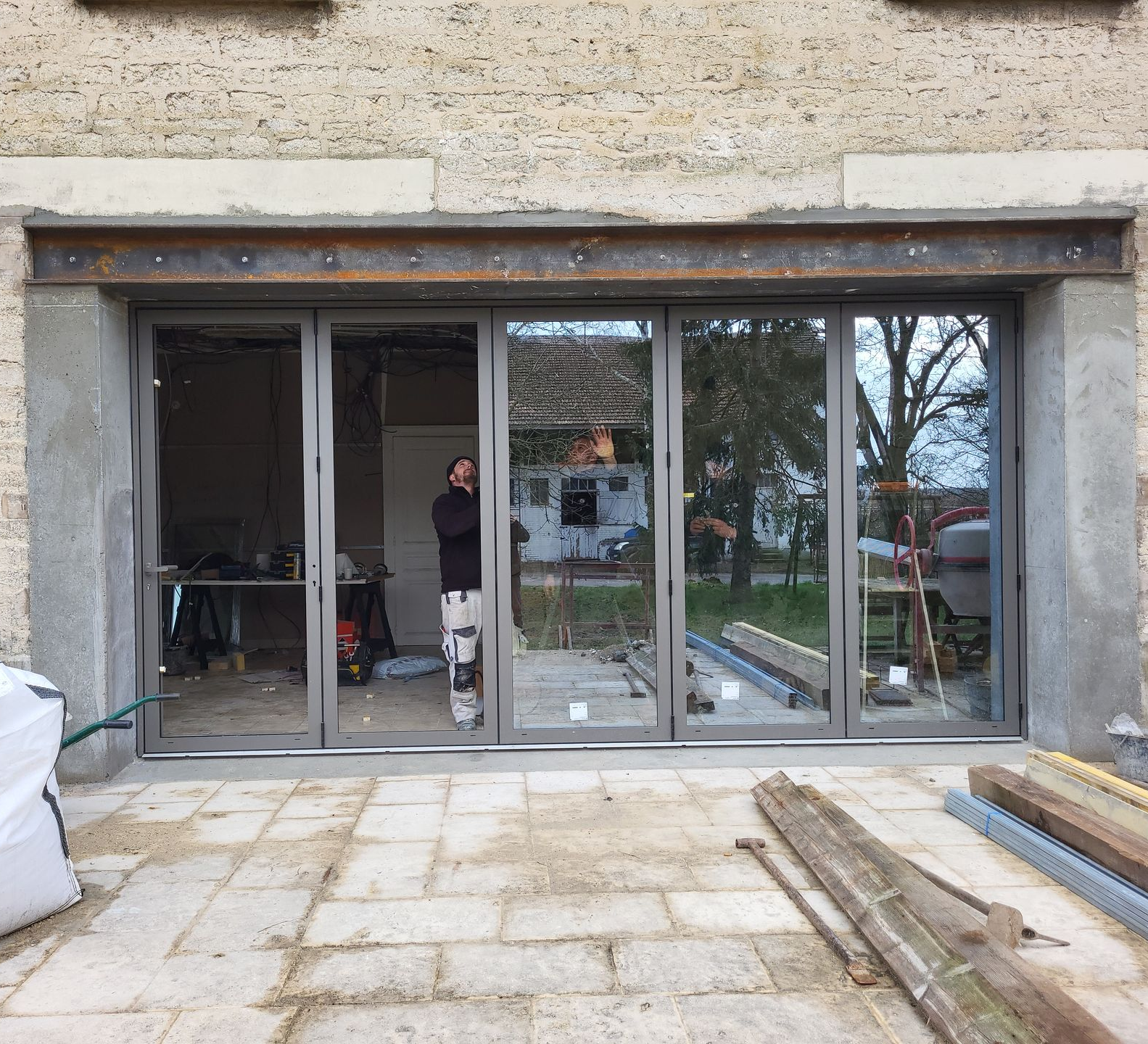Un homme se tient debout à une fenêtre ouverte, tenant un pinceau et un seau de peinture, à l'intérieur d'une pièce en construction.
