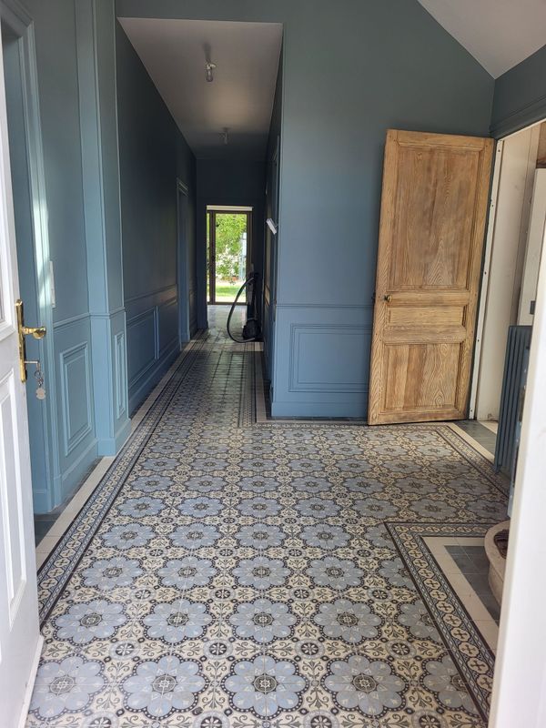 Long couloir avec carrelage à motifs, murs bleus et porte en bois clair.