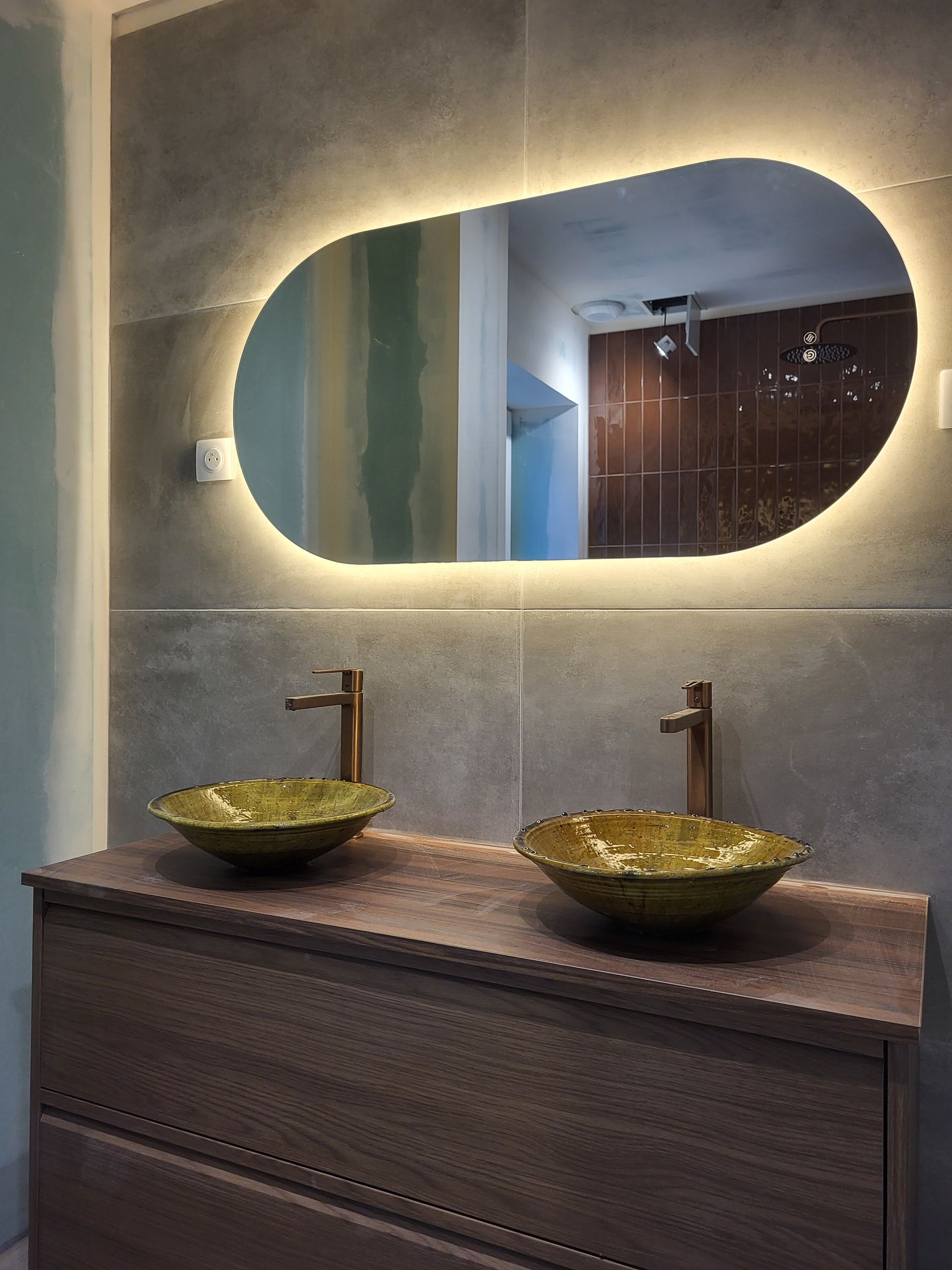 Salle de bains avec miroir ovale éclairé, deux vasques dorées et robinetterie en bronze.