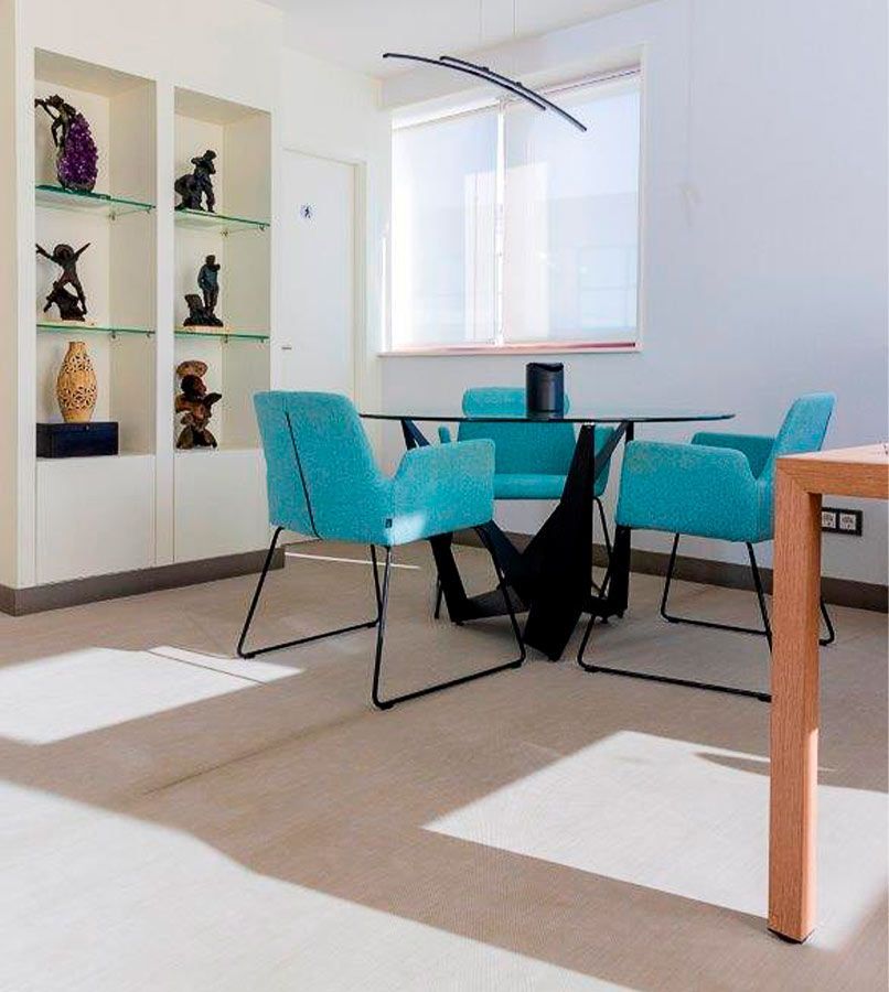 Comedor con sillas turquesas, mesa de cristal y estanterías con obras de arte. Luz natural brillante.