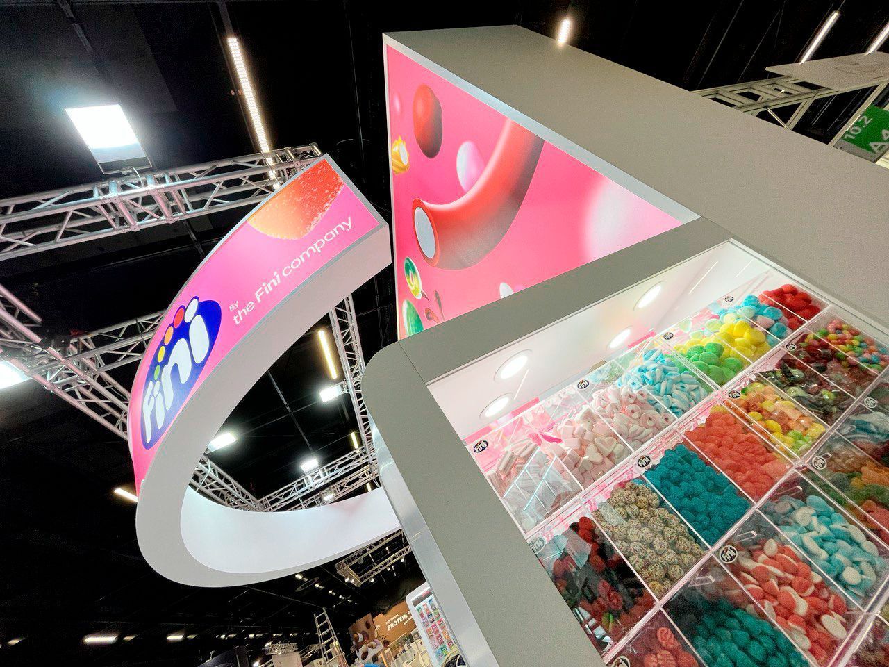 Exhibición de dulces en una feria comercial con un cartel curvo y dulces de colores brillantes en contenedores.