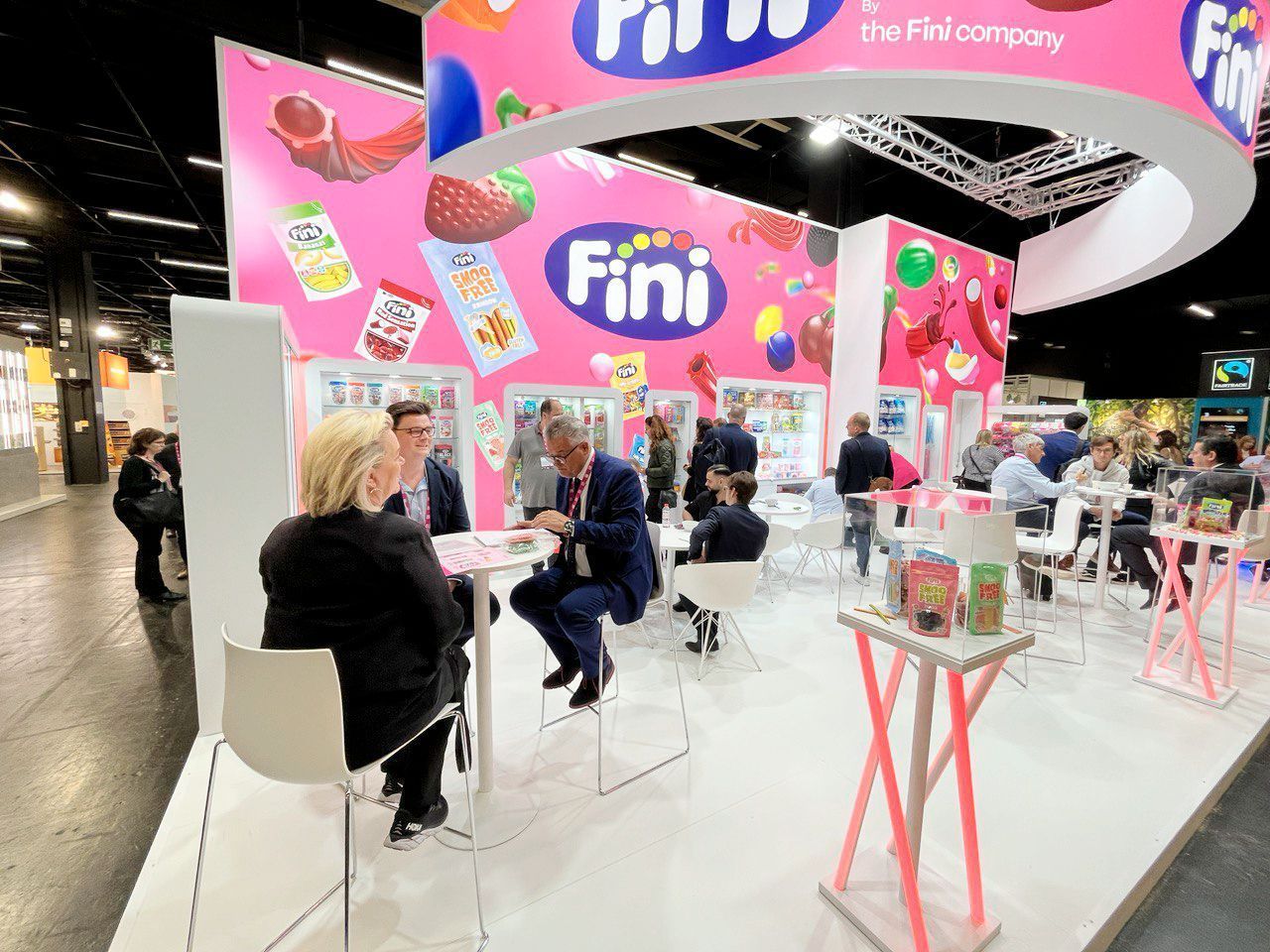 Personas en el stand de dulces Fini en una feria comercial. Letreros rosas y expositores de productos.