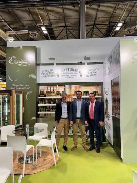 Tres hombres se encuentran dentro de un stand de feria con decoraciones en tonos de aceituna.