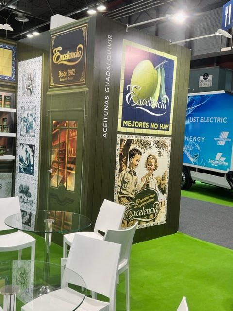 Puesto de aceite de oliva en una feria comercial con carteles. Suelo verde, sillas blancas y mesa de cristal.