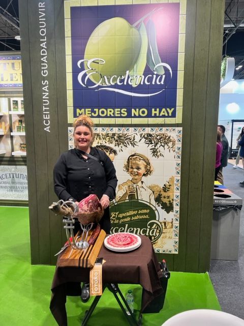 Mujer con jamón en un puesto de aceite de oliva, mostrando el producto con un cartel de estilo vintage.