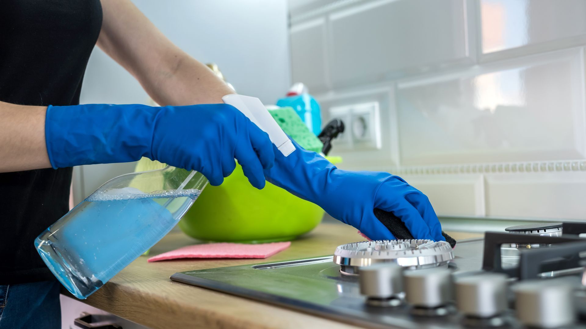 Une personne portant des gants bleus vaporise du produit nettoyant sur une plaque de cuisson tout en tenant une éponge.