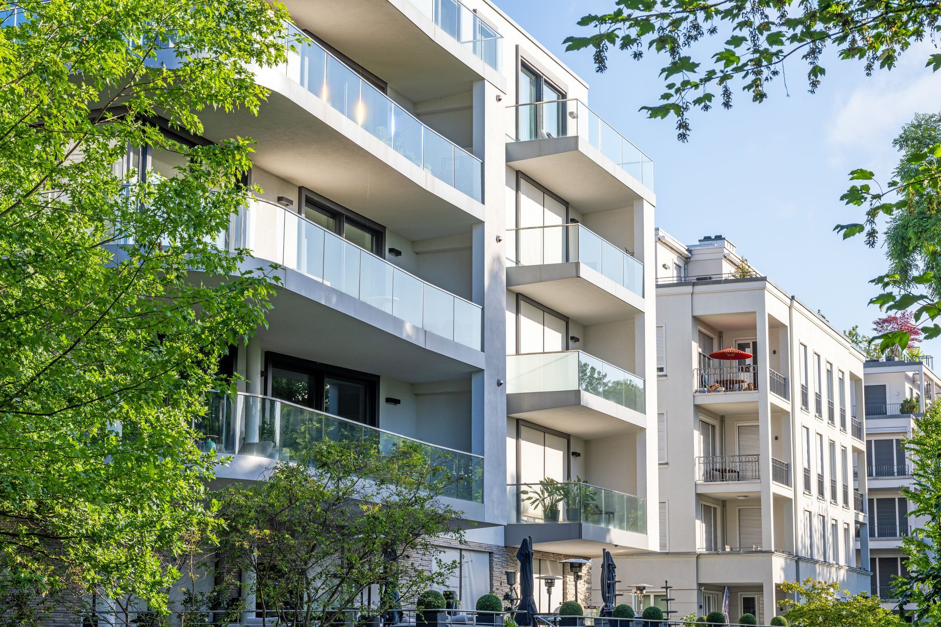 Un immeuble d'appartements moderne, blanc, à plusieurs étages, avec des balcons vitrés, partiellement dissimulés par des arbres verts.