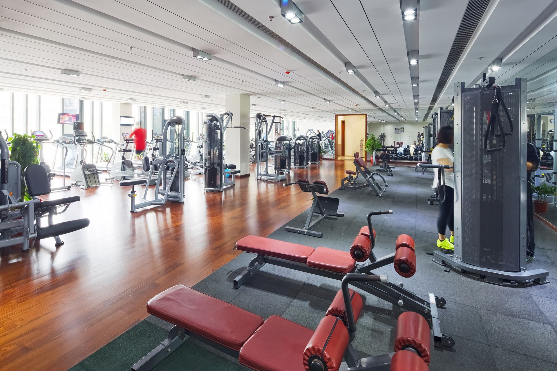 Une salle de sport spacieuse et lumineuse, équipée de rangées d'appareils de musculation, de bancs de musculation rouges et d'un parquet.
