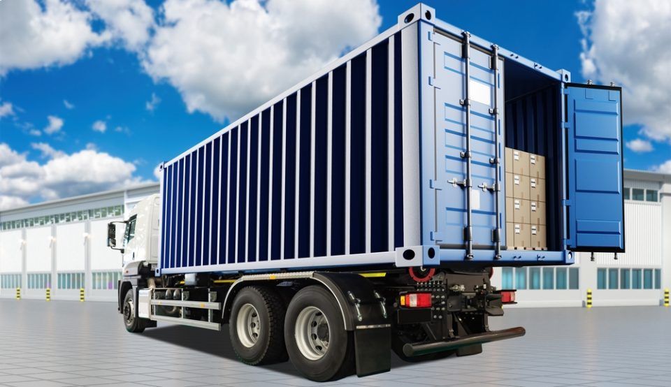 Ein LKW mit einem Frachtcontainer auf der Ladefläche steht vor einem Gebäude.