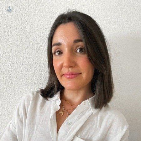 Una mujer que lleva una camisa blanca y un collar está parada frente a una pared blanca.